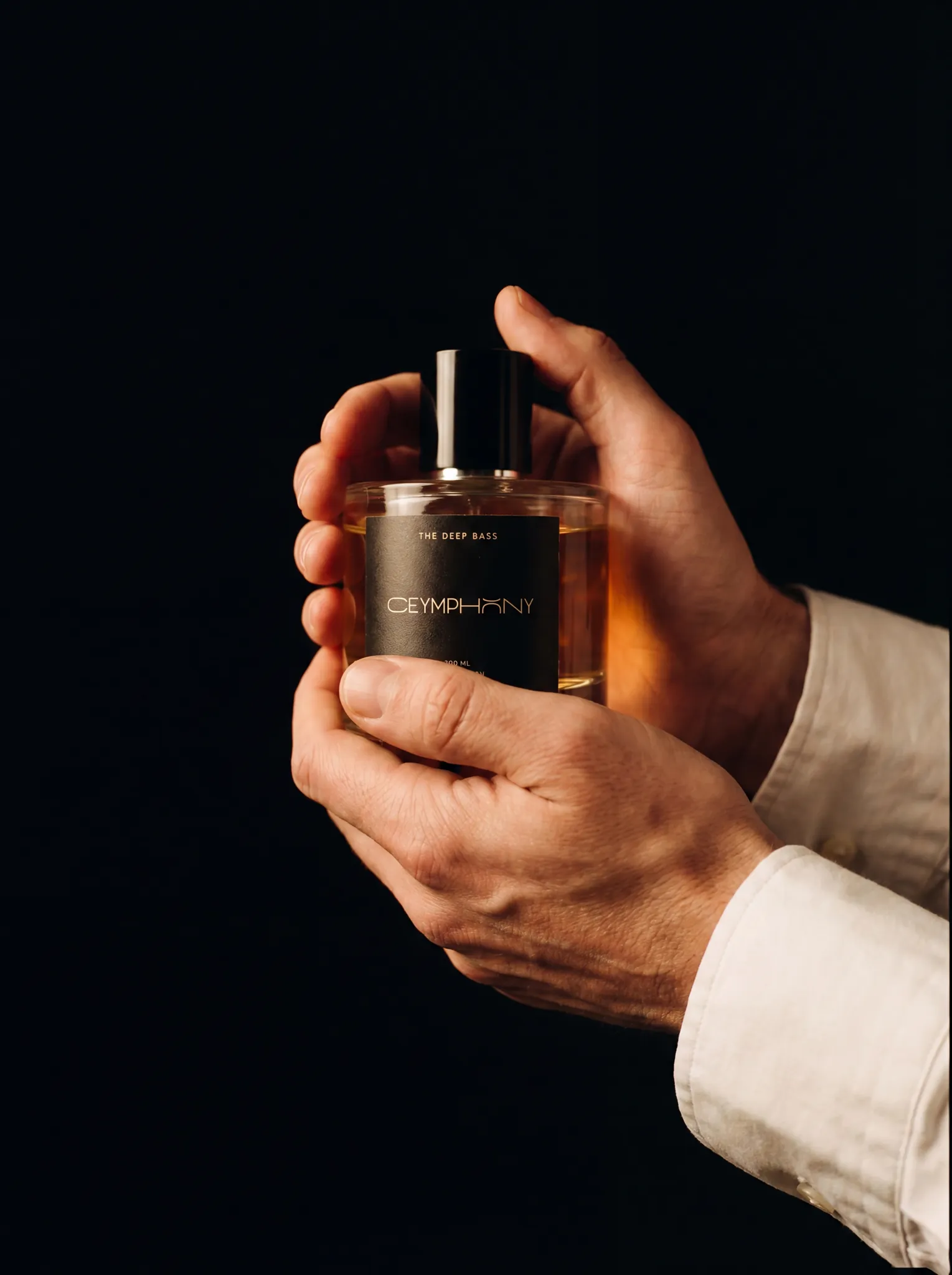 Hands cradling The Deep Bass bottle in a dark interior at a late hour, white cotton cuff, no jewelry.