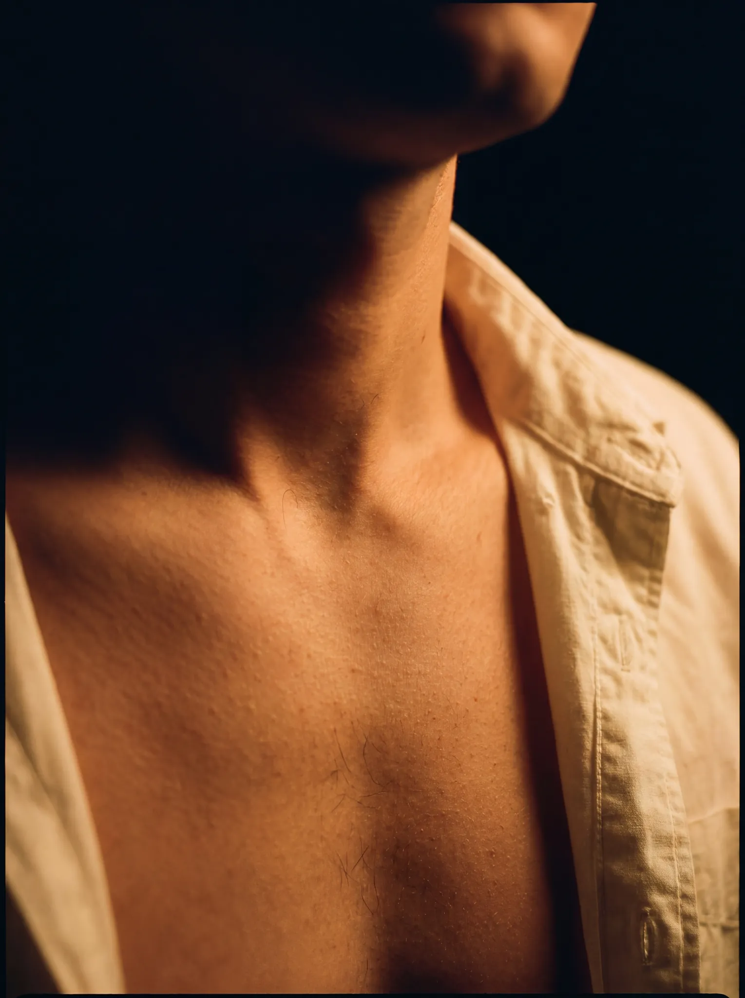 Scent suggestion — collarbone of a figure in an open-collar white cotton shirt, single warm tungsten rim from the right.