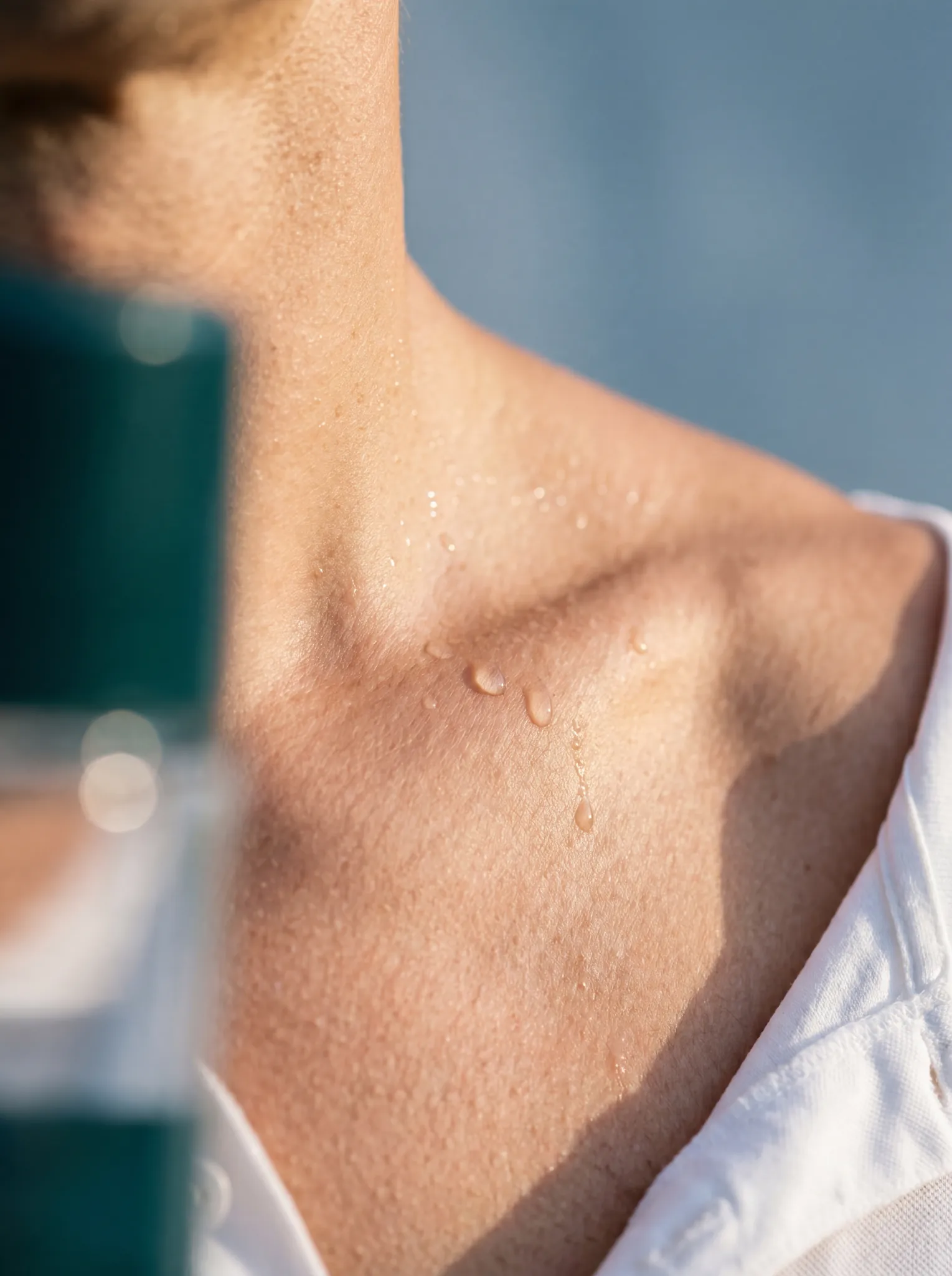 Macro detail — sweat beads on the collarbone of an athletic figure, white open polo, the Melody bottle in deep bokeh at the left edge.